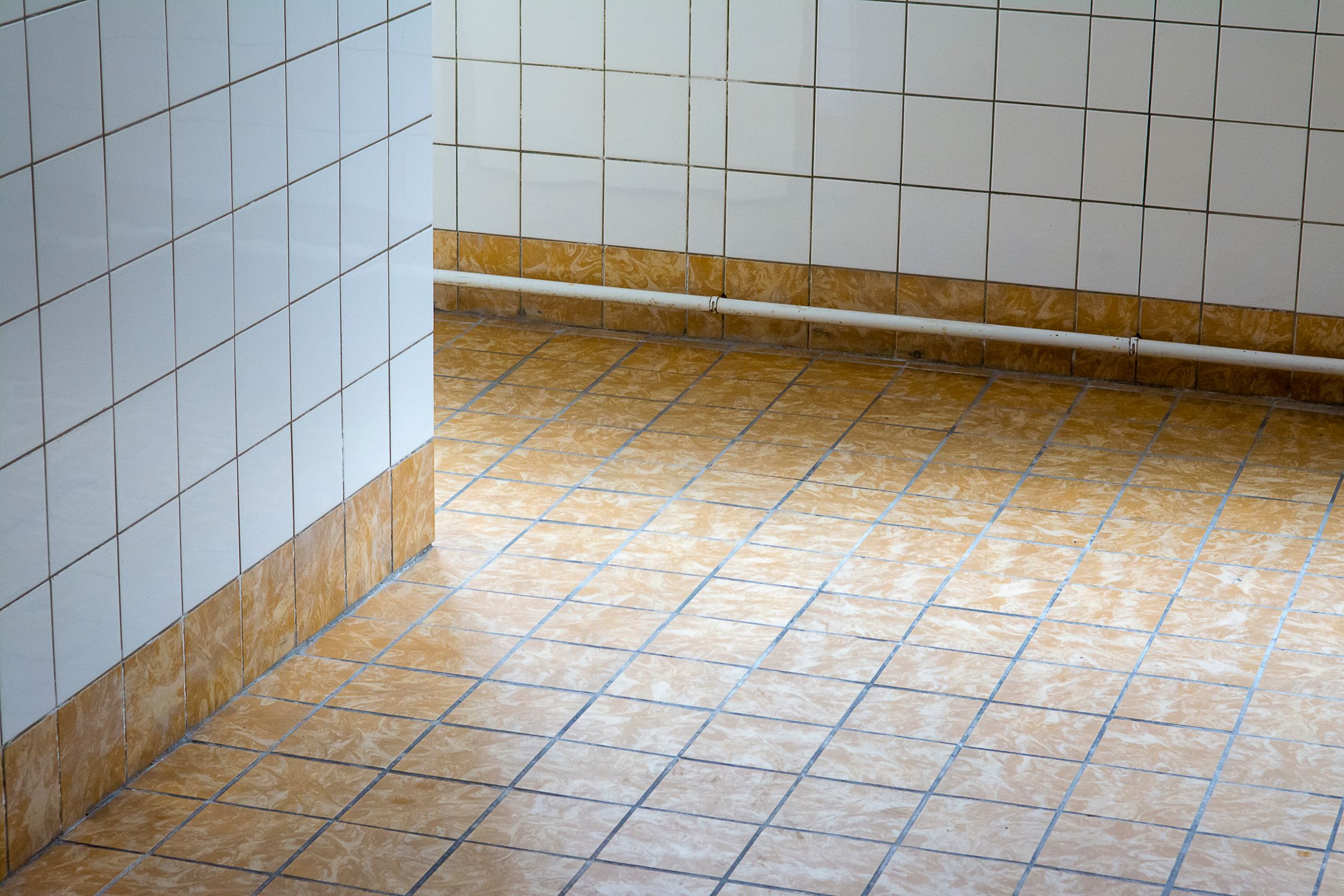 Vanhan märkätilan laatoitus, jossa valkoiset seinälaatat, kuluneet keltaiset lattialaatat ja pinta-asennettu putki.